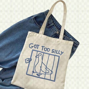 May include: A cream-colored tote bag with a blue illustration of a bird in a cage and the text "GOT TOO SILLY". The bag has two handles and is resting on a pair of blue jeans.