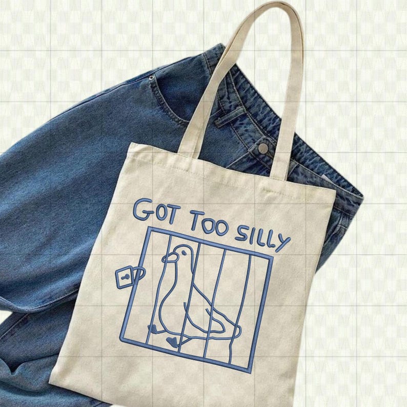 May include: A cream-colored tote bag with a blue illustration of a bird in a cage and the text "GOT TOO SILLY". The bag has two handles and is resting on a pair of blue jeans.