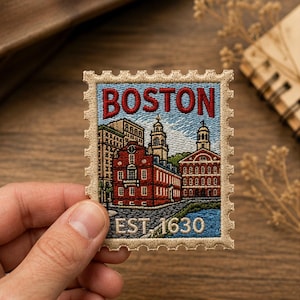 May include: A rectangular embroidered patch shaped like a postage stamp with a beige scalloped border. The design features the red text BOSTON at the top and EST. 1630 at the bottom, with a detailed stitched scene of red brick buildings.