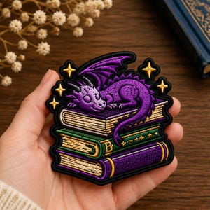May include: An embroidered patch featuring a purple dragon sleeping on a stack of three books. The books are coloured purple and green with gold accents. The design includes four small gold stars surrounding the dragon, all outlined in black thread.