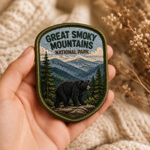 May include: An embroidered patch with an olive green border featuring a black bear standing on a rock in a forest. The background shows blue mountain ranges and clouds under the text GREAT SMOKY MOUNTAINS NATIONAL PARK in dark green thread.