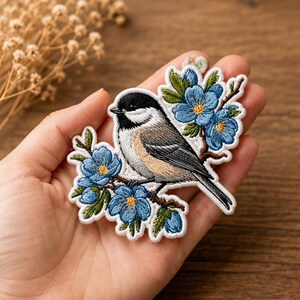 May include: An embroidered patch featuring a black-capped chickadee perched on a brown branch with bright blue flowers and green leaves. The bird has a black head, white cheeks, and grey wings with a light tan underside, all outlined in white thread.