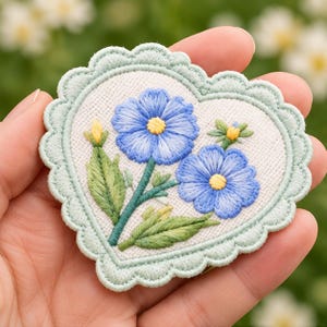 May include: A heart-shaped embroidered patch featuring two blue flowers with yellow centers, green leaves, and buds on a white fabric background, finished with a scalloped light green stitched border edge.