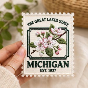 May include: A rectangular embroidered patch shaped like a postage stamp with a cream background. It features a dark green border, the text THE GREAT LAKES STATE, MICHIGAN, and EST. 1837, and a central illustration of pink and white apple blossoms.