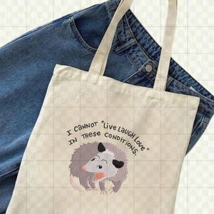 Peut inclure: Un sac fourre-tout cr&egrave;me avec le texte "I CANNOT IN THESE CONDITIONS. Live Laugh Love" et une illustration de dessin anim&eacute; d'un opossum. Le sac a de longues poign&eacute;es beiges et est pos&eacute; sur un fond de jean bleu.