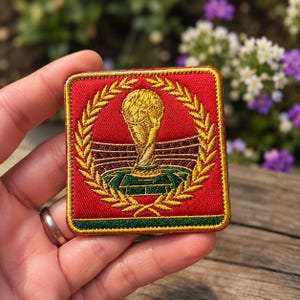 May include: A square, red embroidered patch featuring a gold trophy, surrounded by a gold laurel wreath. The patch has a green and gold border and is likely a sports-related item.