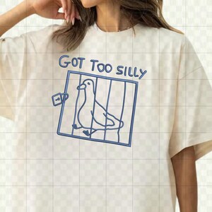 May include: Cream-colored t-shirt with a blue outline drawing of a bird in a cage. The text "GOT TOO SILLY" is above the cage in blue. The shirt has short sleeves and a crew neck.