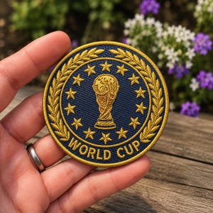 May include: Embroidered patch with a navy blue background and gold accents. The patch features a gold trophy, stars, and the words "WORLD CUP" in a circular design. The patch is round and has a gold border.