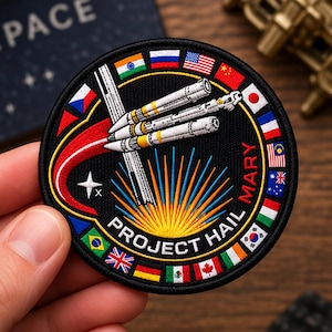 May include: A circular embroidered patch featuring a black background with a white rocket ship launching upwards. The text "PROJECT HAIL MARY" is arched at the bottom. A sunburst design sits below the rocket, surrounded by a border of various national flags.