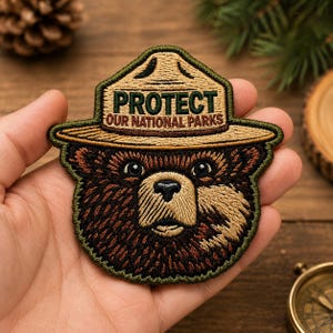 May include: An embroidered patch featuring a brown bear wearing a tan ranger hat. The hat displays the text PROTECT OUR NATIONAL PARKS in dark green and brown lettering. The patch has a dark green border and detailed stitching throughout.