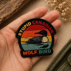 May include: A circular embroidered patch featuring a black and white loon swimming on water against a sunset background of red, orange, and yellow stripes. The text STUPID CANADIAN is arched at the top and WOLF BIRD is at the bottom in bold letters.