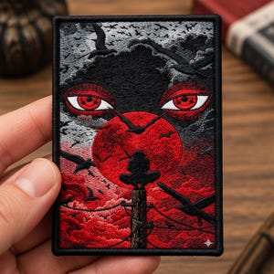 May include: A rectangular embroidered patch featuring a dark, atmospheric scene with a large red moon, a silhouette of a figure perched on a utility pole, and two prominent red eyes above. The design includes black birds, clouds, and a black border.