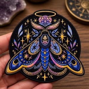 May include: A circular embroidered patch featuring a moth with blue, purple, and gold thread. The moth has a golden halo above its head, intricate wing patterns with crescent moons, and surrounding star motifs set against a solid black background.