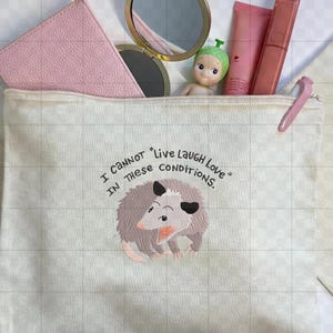 Peut inclure: Une trousse de maquillage en toile avec une fermeture &eacute;clair, orn&eacute;e d'un opossum de dessin anim&eacute; et du texte "I cannot 'Live Laugh Love' in these conditions." Le sac contient des articles de maquillage, dont un portefeuille rose, un miroir et des produits pour les l&egrave;vres.