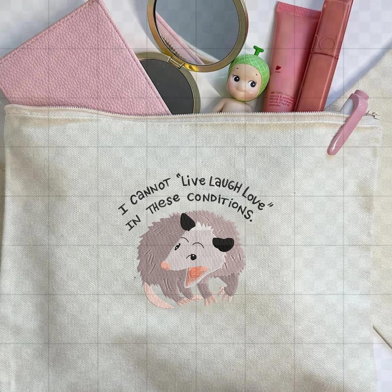 Peut inclure: Une trousse de maquillage en toile avec une fermeture &eacute;clair, orn&eacute;e d'un opossum de dessin anim&eacute; et du texte "I cannot 'Live Laugh Love' in these conditions." Le sac contient des articles de maquillage, dont un portefeuille rose, un miroir et des produits pour les l&egrave;vres.