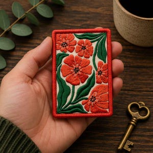 May include: A rectangular embroidered patch featuring a cream-colored background with vibrant red flowers and deep green leaves. The patch is framed by a thick, stitched red border, showcasing detailed needlework with visible thread textures.