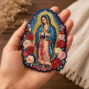 May include: An embroidered patch featuring the Virgin of Guadalupe with a blue mantle, pink robe, and golden rays. The figure is surrounded by red, pink, and cream-colored roses, all set against a dark blue scalloped border with intricate stitching.