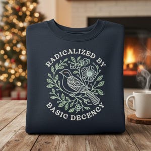 May include: Navy blue sweatshirt with the text "RADICALIZED BY BASIC DECENCY" in white. The design features a bird and floral elements in a circular arrangement. The sweatshirt is displayed on a wooden surface.