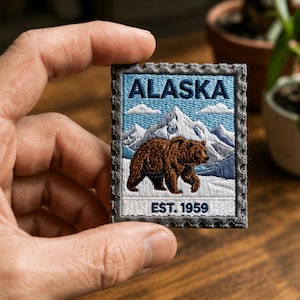 May include: A rectangular embroidered patch shaped like a postage stamp featuring a brown bear walking in front of snow-capped mountains. The design includes the text ALASKA at the top and EST. 1959 at the bottom, with a grey scalloped border.