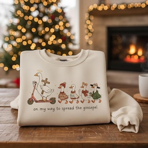 Può includere: Felpa color crema con un'illustrazione stravagante di oche. Un'oca guida un monopattino, tenendo una croce, con la scritta "on my way to spread the goosepel". Altre oche sono vestite per l'inverno. Lo sfondo include un albero di Natale e un camino.