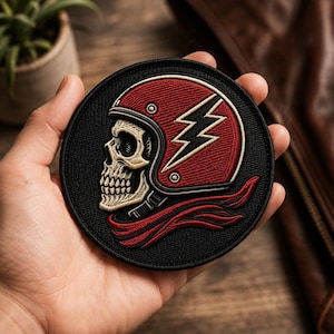 May include: A circular embroidered patch featuring a beige skull wearing a red motorcycle helmet with a white lightning bolt emblem on the side. The design includes a red scarf flowing beneath the helmet, all set against a solid black background.