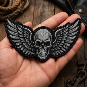 May include: An embroidered patch featuring a central human skull with detailed shading in shades of grey and black. Large, feathered wings extend outwards from both sides of the skull. The entire design is outlined with a thick black border.