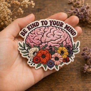 May include: An embroidered patch featuring a pink brain illustration with a colorful floral arrangement at the base. The text BE KIND TO YOUR MIND is stitched in dark maroon thread along the top edge of the off-white fabric backing.