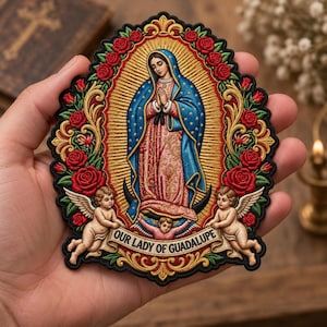 May include: An embroidered patch featuring Our Lady of Guadalupe in a blue mantle and pink dress, surrounded by red roses, gold rays, and two winged cherubs. A banner at the bottom displays the text OUR LADY OF GUADALUPE in black lettering.