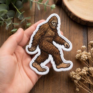 May include: An embroidered patch featuring a brown Bigfoot figure in a walking pose. The creature has dark brown fur, lighter tan skin on the hands and feet, and a white border outline. The design is detailed with stitched textures throughout.