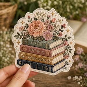 Może przedstawiać: Haftowana naszywka przedstawiająca stos czterech książek z napisem "LET'S GET LOST IN A GOOD BOOK" na grzbietach. Książki zwieńczone są bukietem różowych i brzoskwiniowych kwiatów, zielonymi liśćmi oraz małymi złotymi gwiazdkami na kremowym tle.