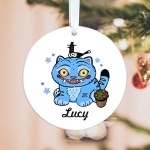 May include: A round, white Christmas ornament featuring a cartoon blue cat with large yellow eyes, a black tail, and a small flower pot. The cat wears a hat and holds a bottle. The name "Lucy" is written below the cat.