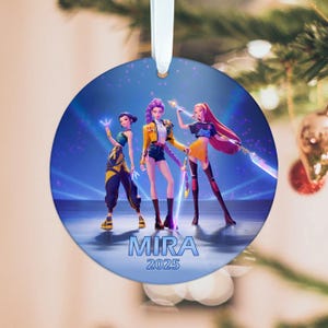 Op de afbeelding: Ronde kerstversiering met drie geanimeerde figuren in stijlvolle outfits. De versiering heeft een blauwe achtergrond met het woord "MIRA" en het jaar "2025" erop gedrukt. Een wit lint is bevestigd om op te hangen.