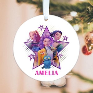 Op de afbeelding: Een witte, ronde kerstversiering met een cartoonillustratie van drie vrouwen en een blauwe kat. De versiering heeft een paars sterontwerp en de naam "AMELIA" in roze. Een wit lint is bevestigd om op te hangen.