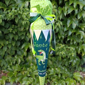 Könnte beinhalten: Eine grüne und weiße Schultüte mit einem Dinosaurier-Design. Die Tüte ist mit einem grün-weißen Sternmuster, grünen Bändern und dem Namen "Jonte" verziert. Der Text "Schulkind 16.08.2023" ist ebenfalls sichtbar.