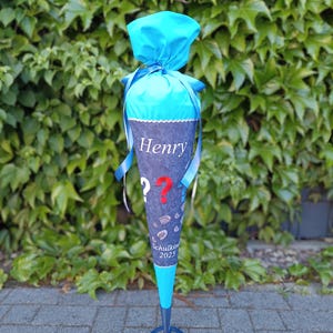 Könnte beinhalten: Eine blaue und graue Schultüte mit dem Namen "Henry" in weißer Schrift. Die Tüte hat einen blauen Deckel, ein Fragezeichen und den Text "Schulkin 2025". Bänder und ein dekorativer Fuß runden das Design ab.