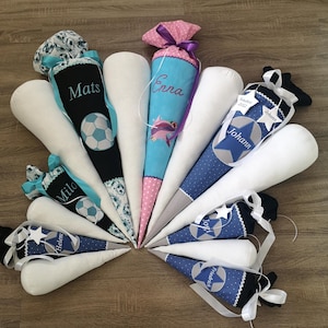 Könnte beinhalten: Eine Sammlung von 10 bunten Schultüten mit verschiedenen Designs und bestickten Namen. Die Schultüten sind in einem Kreis auf einer Holzoberfläche angeordnet. Die Namen auf den Schultüten sind Mats, Enna, Milo, Helen, Johann und Jogi.
