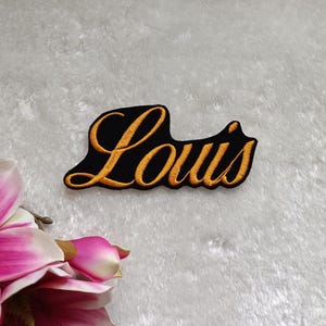 Könnte beinhalten: Ein schwarzer, gestickter Aufnäher mit dem Namen "Louis" in goldener Schrift. Der Aufnäher hat einen schwarzen Rand und ist vor einem weißen, strukturierten Hintergrund platziert. Eine rosa-weiße Blume befindet sich in der unteren linken Ecke.