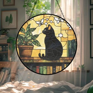 May include: Round stained glass window featuring a black cat silhouette with orange ear tips, sitting on a shelf with books. The background is yellow with butterflies and stars. A potted plant is on the left. The window is hanging from a chain.