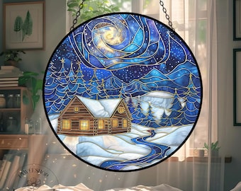 Op de afbeelding: Rond gebrandschilderd glazen raamkunst met een winterhutscène. Het kunstwerk toont een hut met verlichte ramen, een besneeuwd landschap en een sterrenhemel in blauw- en goudtinten. Het stuk wordt opgehangen door een zwarte ketting.