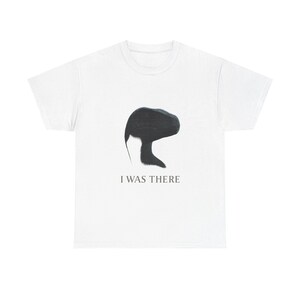 Peut inclure: Un t-shirt blanc à manches courtes arborant une silhouette noire de tête et de cou de dinosaure au centre. Sous le motif, le texte I WAS THERE est imprimé dans une police simple, sombre et en majuscules. Le t-shirt a un col rond et des manches.