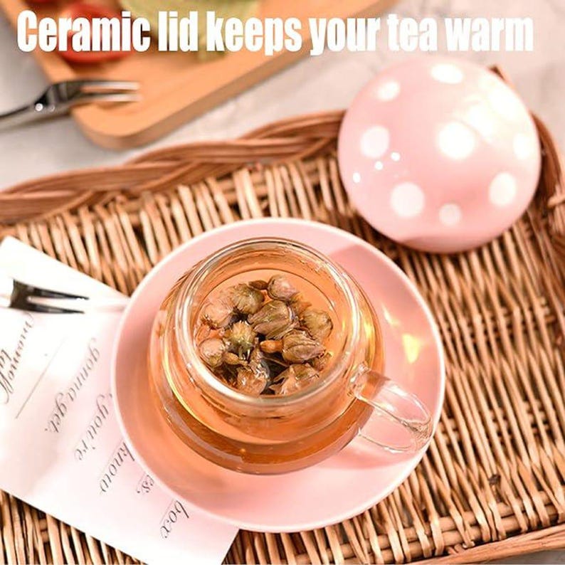 May include: A glass teacup filled with tea and flower buds, resting on a pink saucer. A pink ceramic lid with white polka dots sits nearby. The text "Ceramic lid keeps your tea warm" is at the top.
