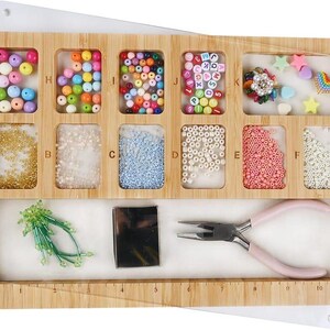 May include: A wooden bead organizer with multiple compartments filled with colorful beads, sequins, and craft supplies. Includes a pair of pink-handled pliers. The organizer is on a white surface.