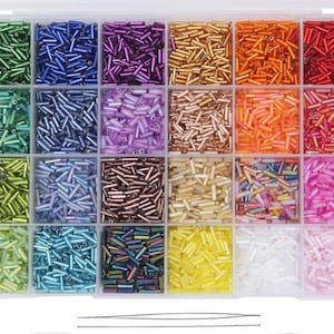 May include: A clear plastic organizer filled with colorful glass seed beads. The beads are in various shades of green, blue, purple, gold, orange, and pink. The organizer has 24 compartments, each filled with a different color of beads. Two beading needles are at the bottom.