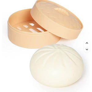 May include: A beige bamboo steamer with an open lid and a white, round bun. The steamer has a perforated base and a textured lid. The bun is smooth and has a spiral design on top. The items are on a white background.