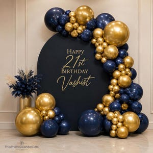 May include: A dark navy blue arched backdrop featuring the gold text Happy 21st Birthday Vashist, decorated with a dense garland of metallic gold and navy blue balloons of varying sizes, accompanied by a vase of dark blue and cream dried foliage.