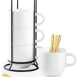 May include: A white ceramic mug set with a black metal stand. The set includes four stacked mugs, three gold spoons, and one additional mug. The mugs have a simple, modern design, perfect for coffee or tea.
