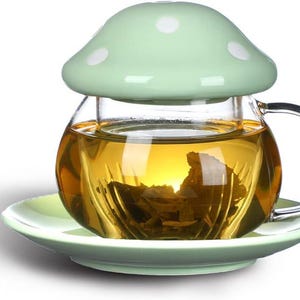 May include: A glass tea cup with a mushroom-shaped lid and saucer. The cup is filled with tea, and the lid and saucer are light green with white polka dots. The cup has a clear handle.