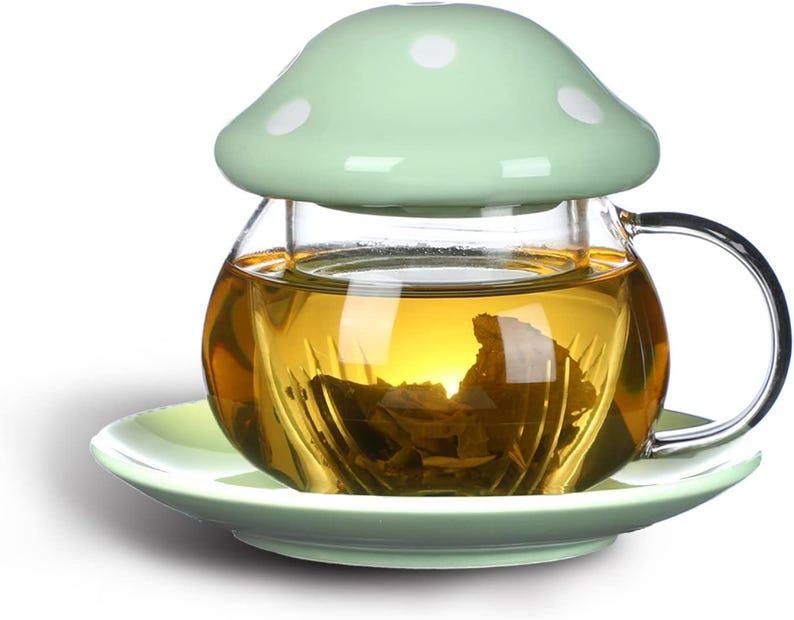 May include: A glass tea cup with a mushroom-shaped lid and saucer. The cup is filled with tea, and the lid and saucer are light green with white polka dots. The cup has a clear handle.