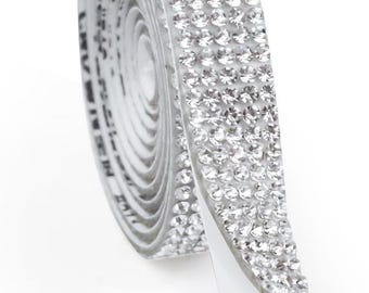 May include: A roll of silver rhinestone ribbon with a self-adhesive backing. The ribbon is covered in small, sparkling rhinestones, creating a shimmering effect. The roll is partially unrolled, revealing the adhesive backing.