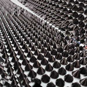 May include: Close-up of a surface covered in rows of black, cone-shaped studs. The studs are evenly spaced and create a textured, geometric pattern. The background is white, providing contrast to the dark studs.